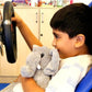 Child in a racing car holding a gray stuffed animal, with a cabinet and various items in the background.
