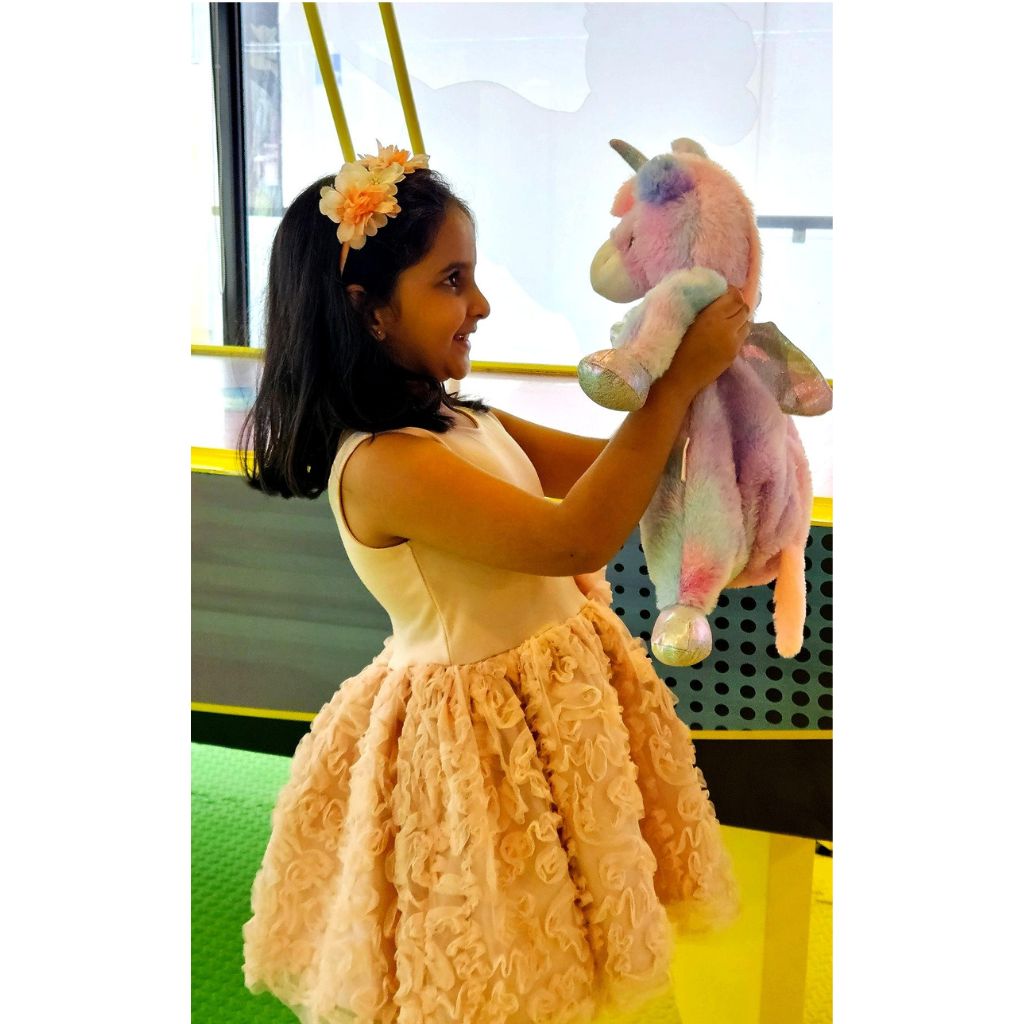 Young girl in a peach dress holding a pink unicorn plush toy on a green floor.